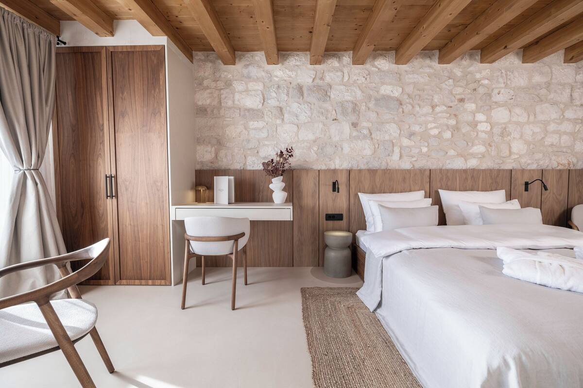 Un dormitorio con pared de piedra, vigas de madera en el techo, una cama, un escritorio y una silla.
