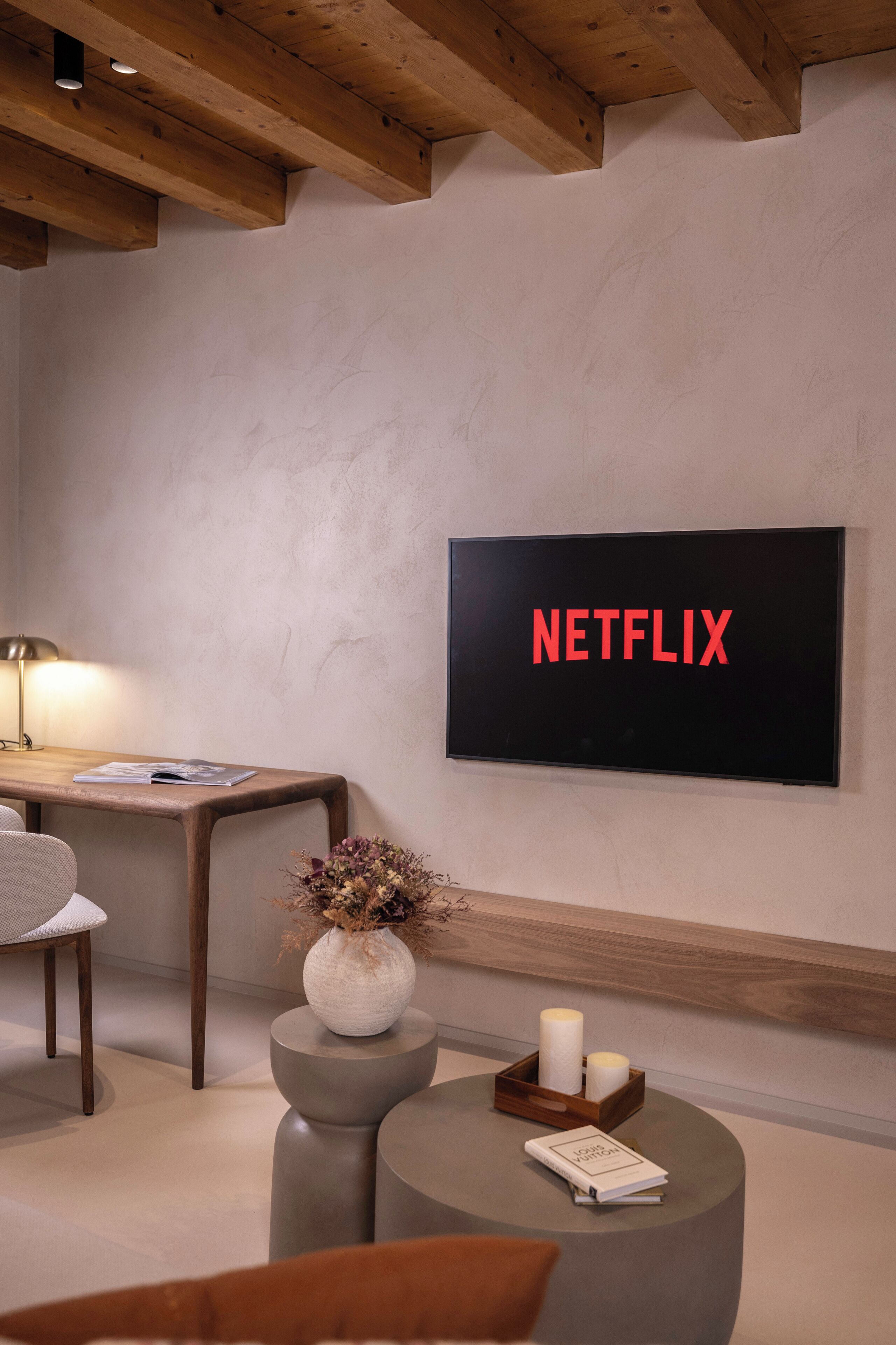 Un salon moderne avec un téléviseur affichant le logo « NETFLIX », un bureau en bois avec une lampe, un vase contenant des fleurs séchées et une petite table avec des bougies et un livre.