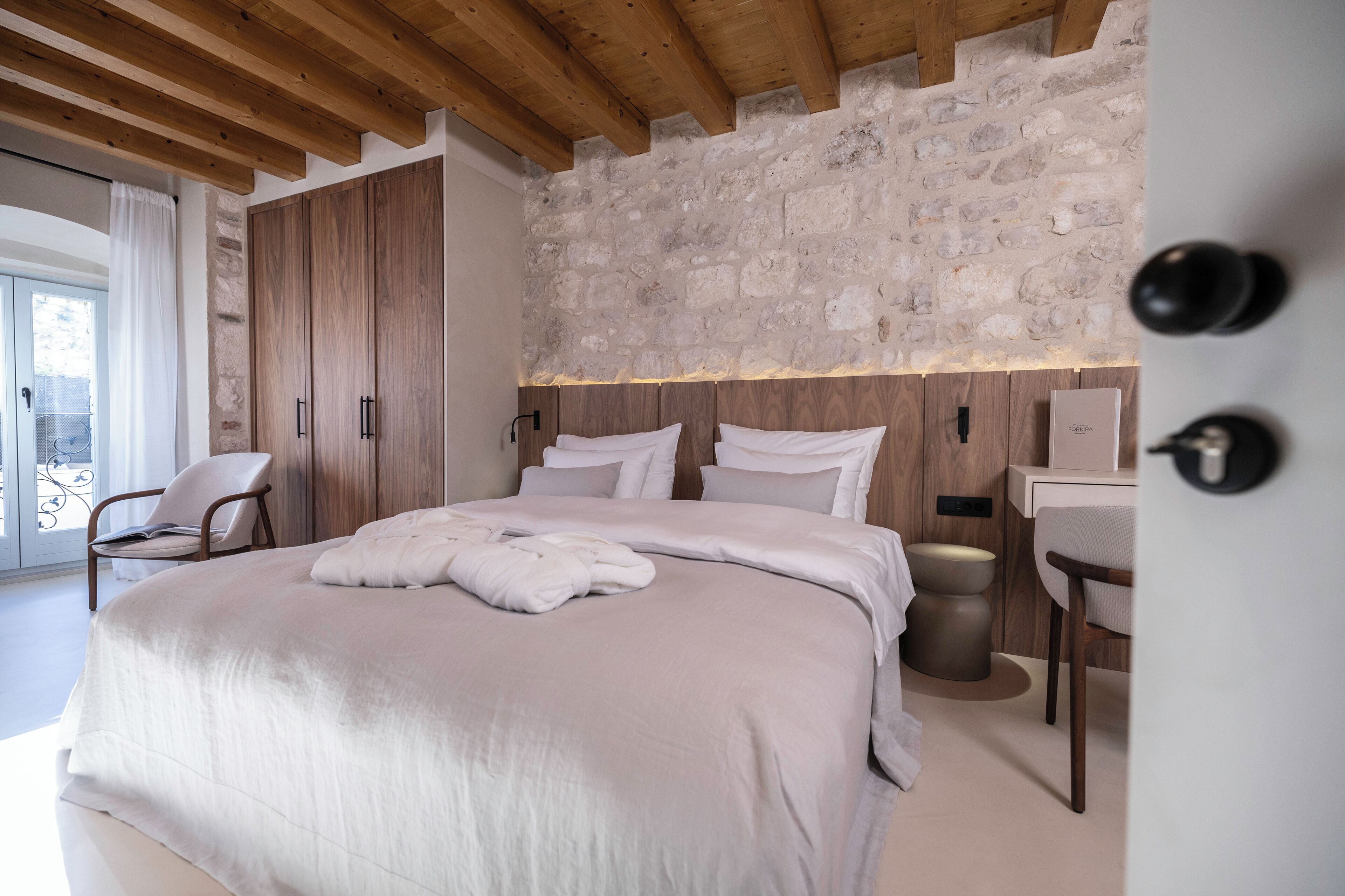 A bedroom with a large bed, wooden wardrobe, stone wall, and a small bathroom with a sink and mirror.