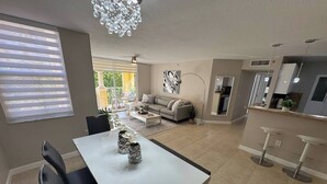 Eine moderne Küche mit einer zentralen Kochinsel, einem Essbereich mit weißem Tisch und schwarzen Stühlen sowie einem Wohnbereich mit Sofa und Couchtisch.