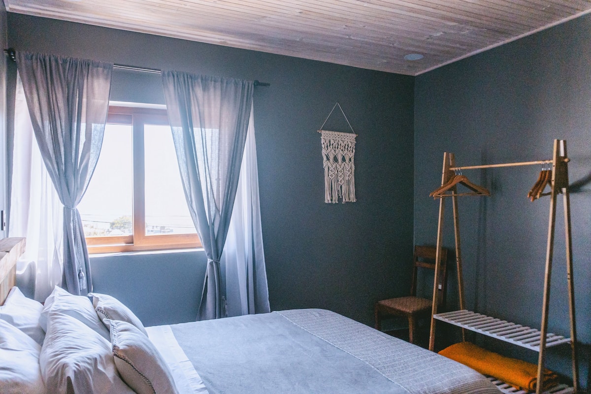 A bedroom with a bed, a window with curtains, a macrame wall hanging, and a wooden drying rack.