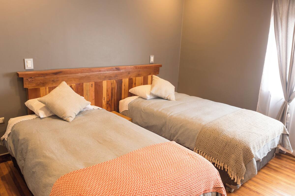 A room with two beds, each with a wooden headboard, grey bedding, and a patterned throw blanket.