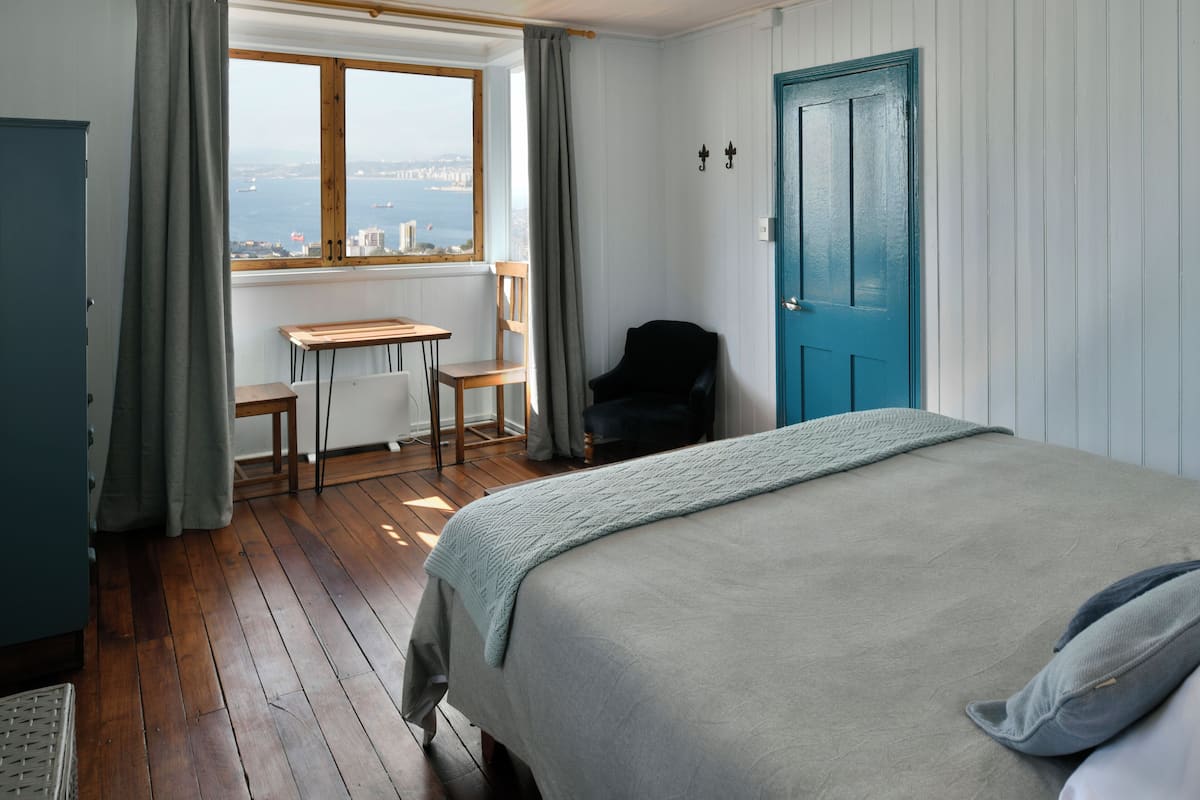 A bedroom with a bed, a desk, a chair, a wardrobe, and a view of the city through a window.