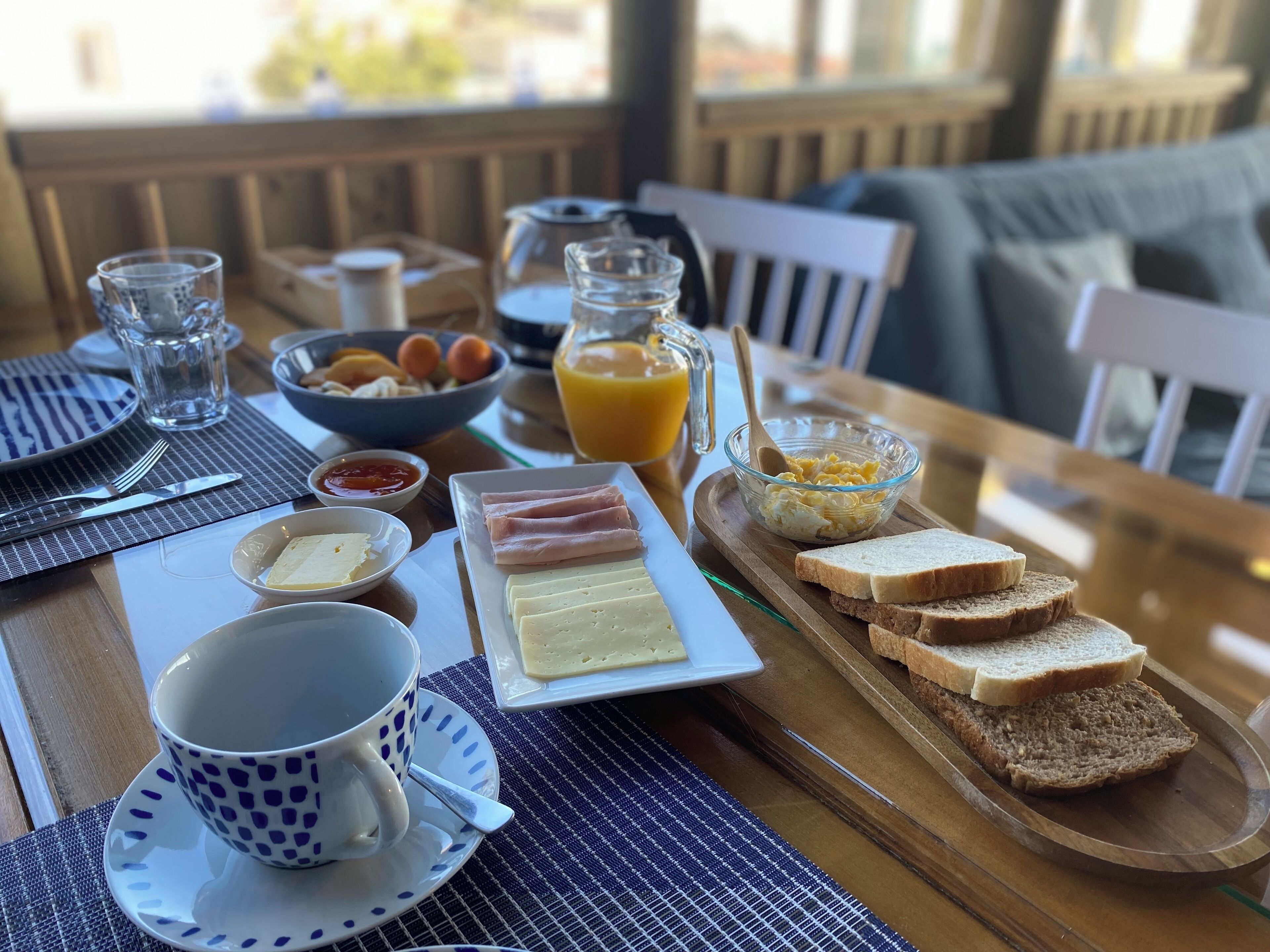 Mesa de café da manhã com pratos de ovos, presunto, queijo, torradas e suco.