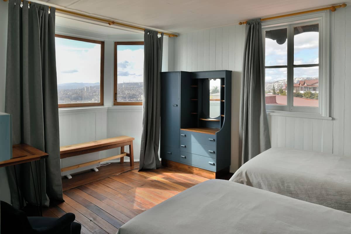 A room with two beds, a bench, a dresser, and a window with a view of a cityscape.