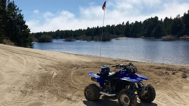 A blue ATV on a sandy beach near a lake, with trees in the background.