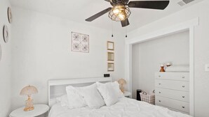 A white bedroom with a bed, a ceiling fan, a bedside table with a lamp, and a dresser. - All American Cottage (Wichita Falls)