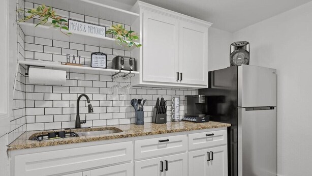 A kitchen with white cabinets, a stainless steel refrigerator, a sink with a faucet, and a countertop with various kitchen items. - All American Lone Star (Wichita Falls)