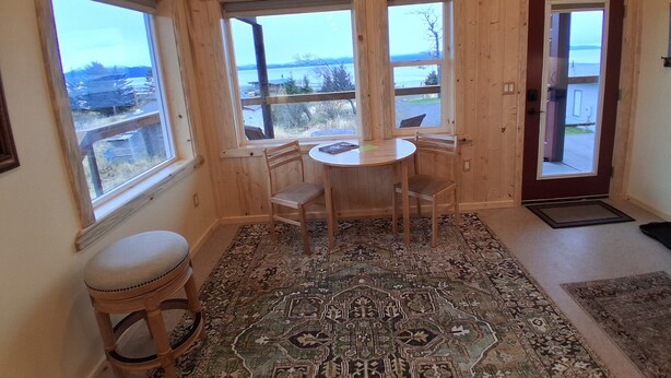 A room with a round wooden table and two chairs, large windows with a view of the outdoors, a wooden door, and a patterned rug on the floor. - GLACIER SUITE, BEACH WALK INN, UPPER LEVEL UNIT
FEATURING HOMER'S FANTASTIC VIEW (Homer)