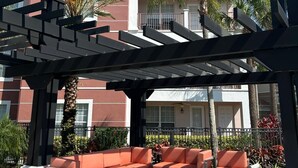 A covered outdoor seating area with orange and grey furniture, palm trees, and a building with balconies in the background. - 4-bedroom apartment in enchanting Orlando with WiFi, AC (Orlando)