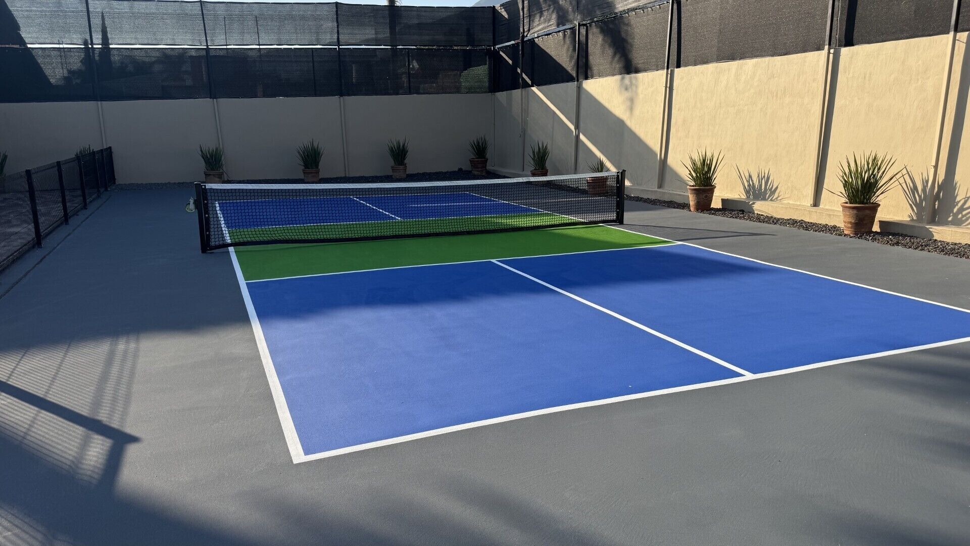 Un court de tennis extérieur, doté d’un filet, entouré de murs et agrémenté de plantes en pot.