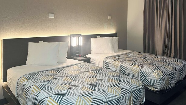 A hotel room with two beds, each with a patterned bedspread, a nightstand with a lamp, and a large window with curtains. - Studio 6 Augusta, Ga (Augusta)