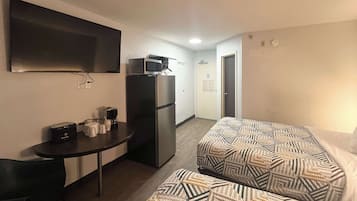 A hotel room with two beds, a small table with a toaster and coffee maker, a refrigerator, a flat-screen TV mounted on the wall, and a door leading to a hallway.