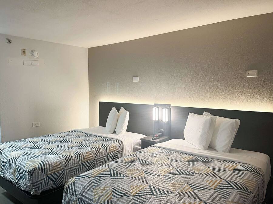 A hotel room with two beds, each with patterned bedding, a nightstand with a lamp, and a wall-mounted light fixture.