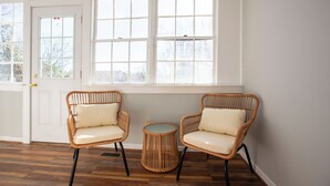 Une pièce comprenant deux fauteuils en osier avec des coussins, une petite table ronde et une grande fenêtre à carreaux.