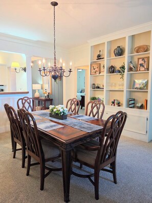 A dining area with a wooden table, chairs, a chandelier, and a bookshelf with various items.