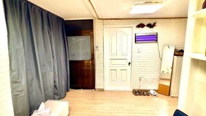 Een compacte kamer met een houten vloer, een witte deur, een klein raam met gordijnen, een spiegel en een wastafel.