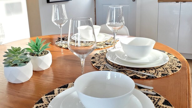 Une table à manger dressée avec de l’assiette blanche, des bols et des verres à vin, entourée de sets de table en fibres tressées et de petites plantes en pot.