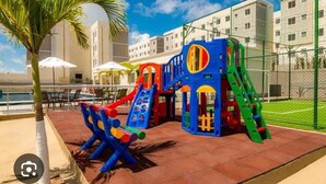 A playground with slides and climbing structures, surrounded by a fence and palm trees.