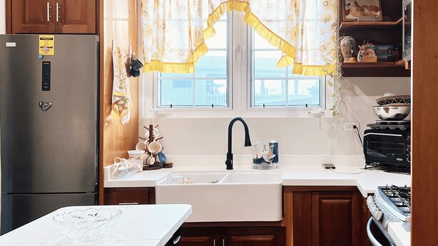 Een keuken met houten kastjes, een roestvrijstalen koelkast, een wit aanrechtblad en een raam met bloemen gordijnen.