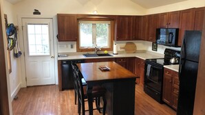 A kitchen with wooden cabinets, a central island with stools, a dishwasher, a stove, and a refrigerator. - Charming Cottage in the Shadow of Day Mountain (Mount Desert)