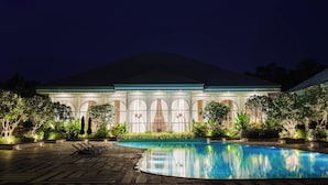 A nighttime view of a hotel with a swimming pool, large arched windows, and lush landscaping.