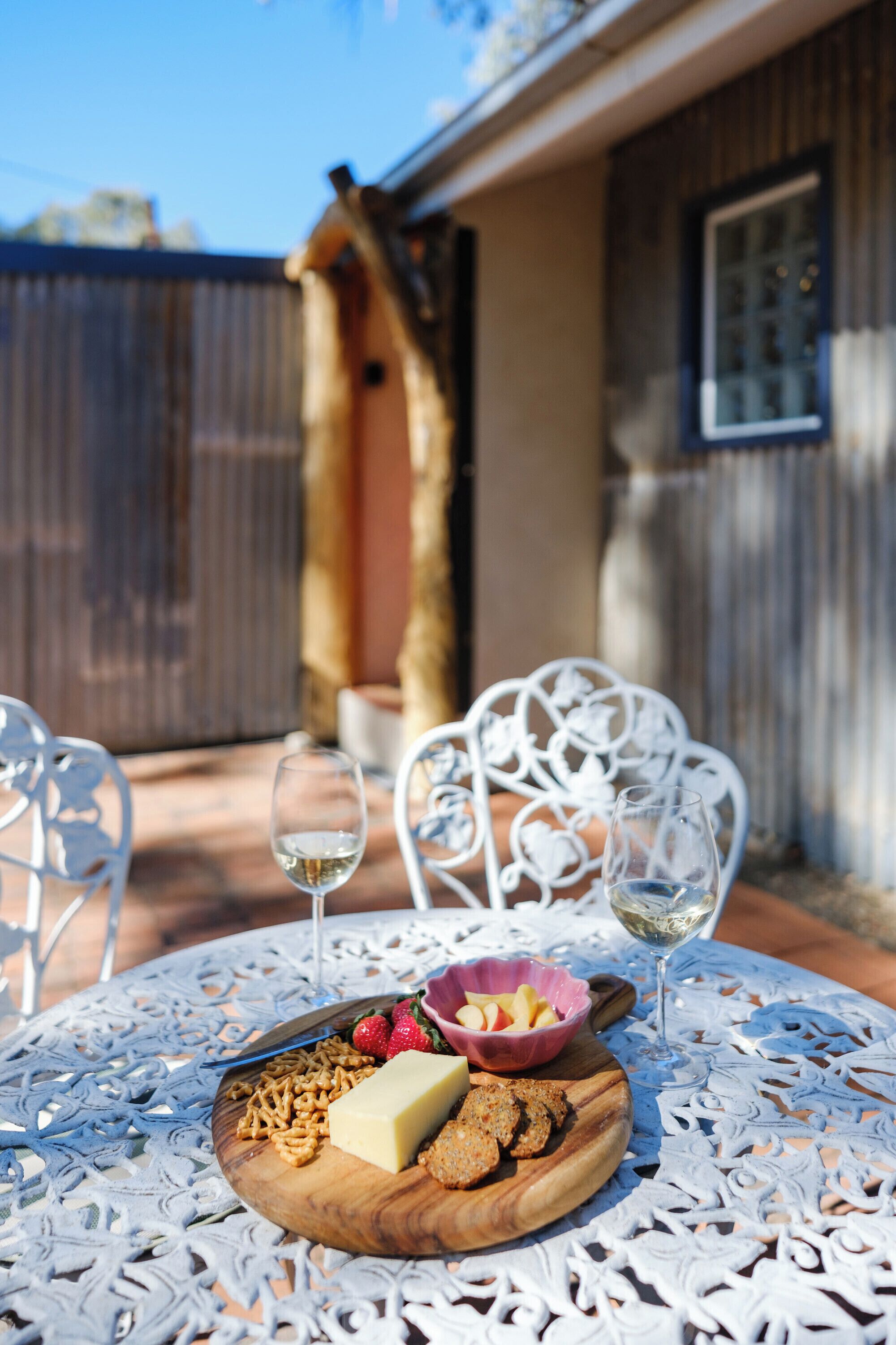 A round table with a white lace tablecloth, a wooden board with cheese, crackers, and fruits, two glasses of white wine, and white chairs.