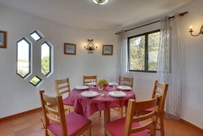 Une salle à manger avec une table dressée pour quatre personnes, des chaises en bois avec des coussins roses, un lustre et une fenêtre avec des rideaux.