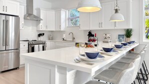 A modern kitchen with white cabinetry, stainless steel appliances, and a central island with blue bowls and plates. - At The Sandbar: Partial Ocean View, Htd Pool, Free Dockage, GameRoom, EV Charger (Islamorada)