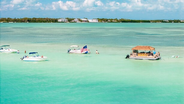 A clear turquoise sea with several boats, including a speedboat with an American flag, anchored near a beach with a thatched-roof structure. - At The Sandbar: Partial Ocean View, Htd Pool, Free Dockage, GameRoom, EV Charger (Islamorada)