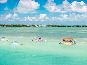 A clear turquoise sea with several boats, including a speedboat with an American flag, anchored near a beach with a thatched-roof structure. - At The Sandbar: Partial Ocean View, Htd Pool, Free Dockage, GameRoom, EV Charger (Islamorada)