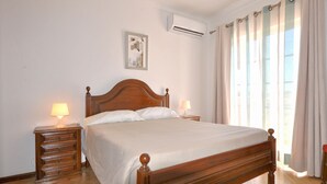 Une chambre à coucher avec un lit en bois, deux tables de chevet avec des lampes, un climatiseur fixé au mur et une fenêtre avec des rideaux.