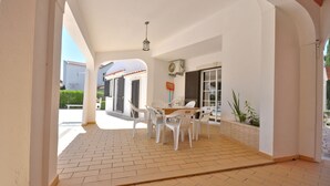 Une terrasse extérieure avec une table et des chaises, un ventilateur de plafond et une vue sur un bâtiment en arrière-plan.