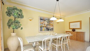 Uma sala de jantar com uma mesa de mármore, cadeiras brancas, um vaso grande e um quadro de barcos.