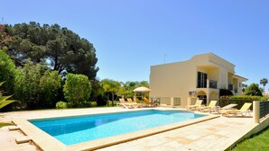Uma piscina com espreguiçadeiras, um edifício com varanda e árvores ao fundo.