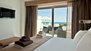 A hotel room with a bed, a TV, and a view of the sea through large windows.