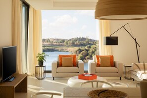 Una sala de estar moderna con un amplio ventanal que ofrece una vista panorámica de un lago y colinas. Hay dos sofás con cojines anaranjados, una mesa de centro y una lámpara de pie.