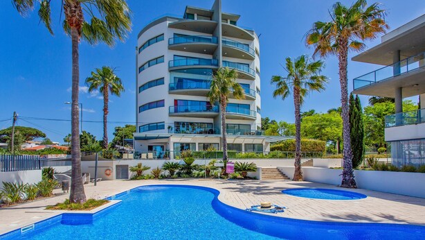 Ein modernes Hotel mit einem zentral gelegenen Pool, umgeben von Palmen und einem mehrstöckigen Gebäude mit Balkonen.