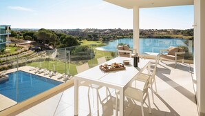 A spacious balcony with a glass railing, a dining table with chairs, a pool, and a view of a golf course and hills.