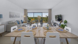 Un comedor moderno con una mesa de madera dispuesta para seis personas, rodeada de sillas blancas. Se ve un sofá y un televisor al fondo, con un amplio ventanal que ofrece vistas a zonas verdes.