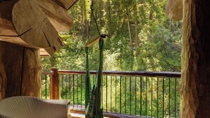 A wooden deck with a hanging bird feeder, a wicker chair, and a view of a forest.