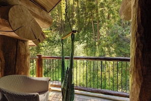 A wooden deck with a hanging bird feeder, a wicker chair, and a view of a forest.
