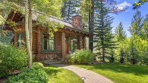 A log cabin with a stone chimney, surrounded by lush greenery and tall trees.