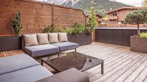 Une terrasse en bois avec un canapé et une table en verre, entourée de jardinières remplies de verdure, offrant une vue sur une chaîne de montagnes.