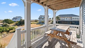 Una terraza de madera con una mesa de picnic, barandillas blancas y vistas a casas y a la playa al fondo.