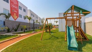 A playground with a slide and swings, a red banner, and a building with windows and palm trees in the background. - Coto Alejandria Mazatlan/2 Albercas/Cerca del Acuario/Cafe/Equipado/Privado (Mazatlán)
