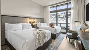 Ein Hotelzimmer mit zwei Betten, einem Schreibtisch, einem Sessel und Blick auf eine verschneite Landschaft.