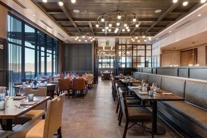 Un restaurant avec de hauts plafonds, des luminaires suspendus et de grandes fenêtres. Les tables sont dressées avec des verres et des couverts.