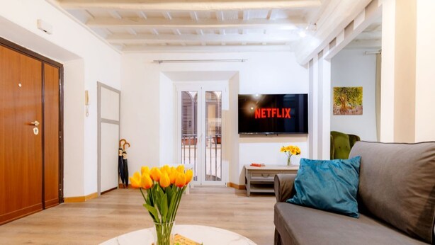 Un salon comprenant un canapé, une table basse ornée d’un vase de fleurs, un téléviseur à écran plat affichant le logo «  NETFLIX », et une porte donnant sur une autre pièce.