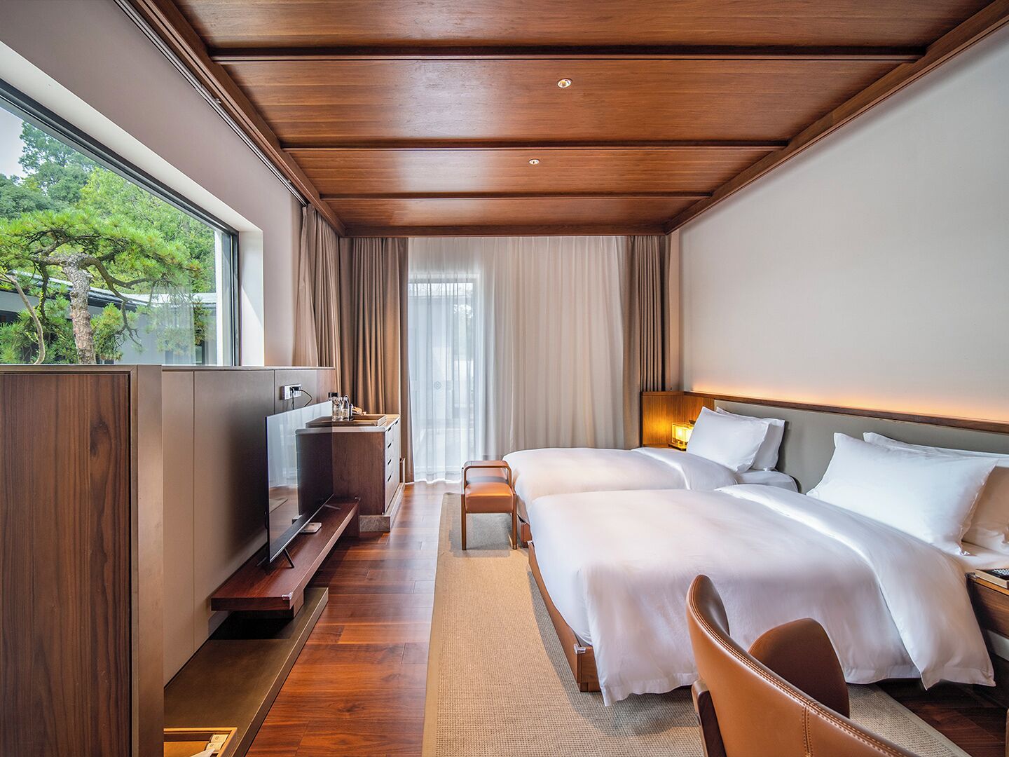 Habitación de hotel moderna con techo de madera, un ventanal con vistas a árboles, dos camas con ropa blanca, una mesita de noche con lámpara y un televisor sobre un mueble de madera.